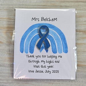 May include: A blue enamel heart-shaped awareness ribbon pin attached to a white card featuring a blue watercolor rainbow illustration. The card text reads: Mrs Belchem, Thank you for helping Me through My highs and lows this year. love Jessie, July 2025.