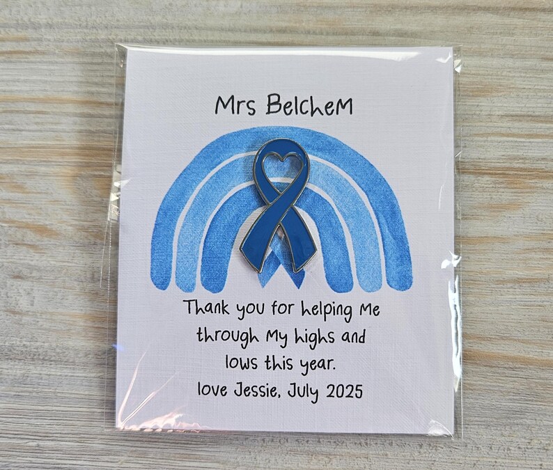 May include: A blue enamel heart-shaped awareness ribbon pin attached to a white card featuring a blue watercolor rainbow illustration. The card text reads: Mrs Belchem, Thank you for helping Me through My highs and lows this year. love Jessie, July 2025.