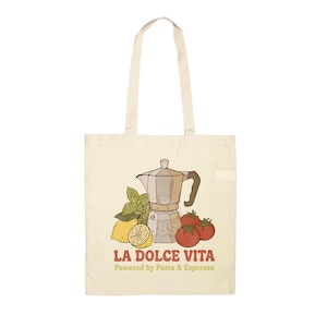 Può includere: Borsa tote color crema con un'illustrazione di caffettiera, pomodori, limone e basilico. Il testo "LA DOLCE VITA" è visualizzato in rosso, con "Powered by Pasta & Espresso" sotto. La borsa ha due manici lunghi.