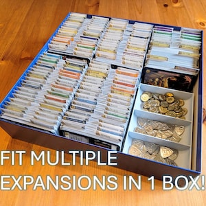 May include: A blue storage box filled with organized game cards and game pieces. The box is divided into compartments, with cards neatly arranged and coins and tokens stored separately. Text on the box reads: FIT MULTIPLE EXPANSIONS IN 1 BOX!