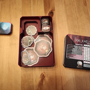 以下が含まれることがあります： 4つの区画を持つ赤いプラスチック製のオーガナイザーには、茶色と金色のデザインの円形トークンや、黒と白のデザインのカードの山など、さまざまなゲームピースが入っています。カードには「006_KAELYFOS SURVIVAL TASK - PERFORMANCE LOG」というテキストが印刷されています。