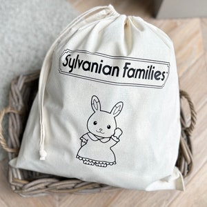 May include: A cream-coloured drawstring bag featuring the text "Sylvanian Families" in a black, rectangular frame. Below is a black and white illustration of a rabbit in a dress, waving.
