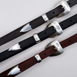 May include: Three belts in dark brown, black, and reddish-brown leather, each featuring a polished silver-tone metal buckle, a matching silver-tone metal keeper, and a pointed silver-tone metal tip. The belts have visible stitching along the edges.