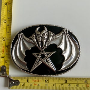 May include: A silver belt buckle with a black enamel background. The buckle features a silver bat with wings spread wide and a pentagram in the center.