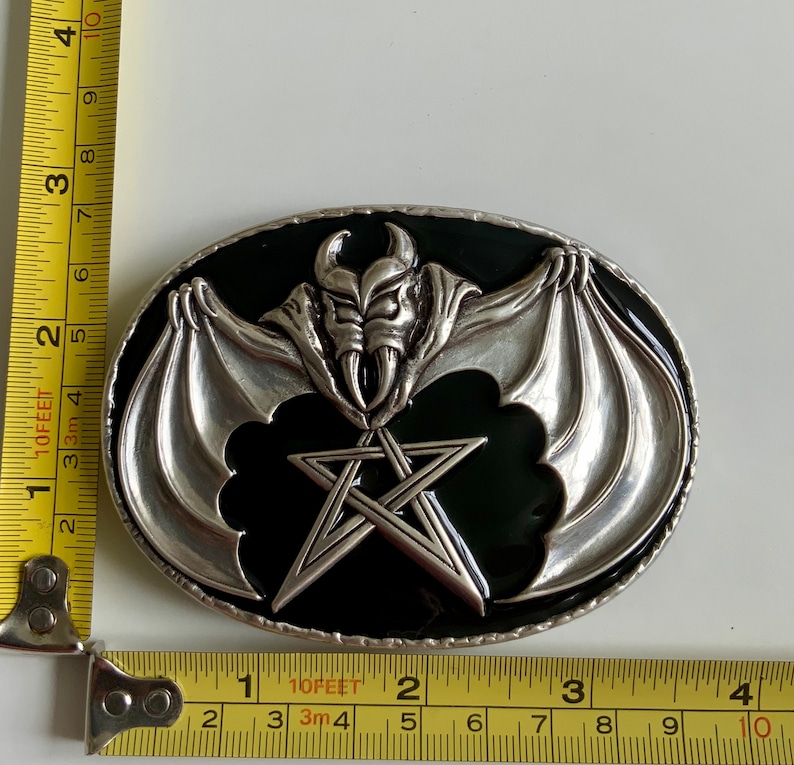 May include: A silver belt buckle with a black enamel background. The buckle features a silver bat with wings spread wide and a pentagram in the center.