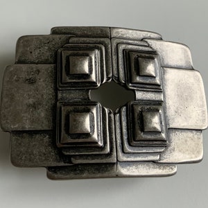 May include: A silver-toned metal belt buckle with a geometric design. The buckle has a square shape with four smaller squares inside, each with a raised center. The buckle has a worn, aged look.