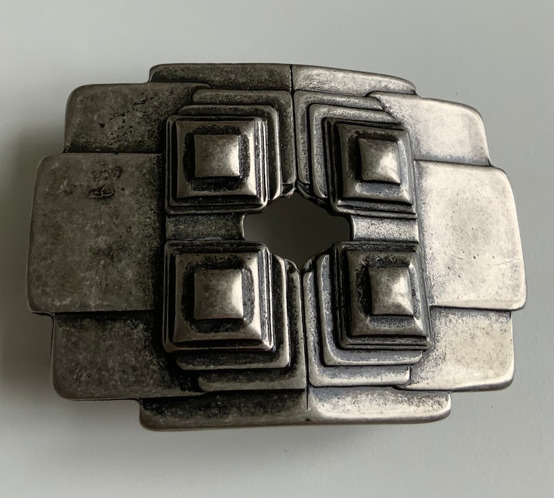 May include: A silver-toned metal belt buckle with a geometric design. The buckle has a square shape with four smaller squares inside, each with a raised center. The buckle has a worn, aged look.