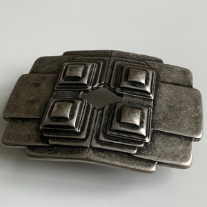 May include: A silver-toned metal belt buckle with a geometric design. The buckle has four square shapes with raised edges and a central hole.