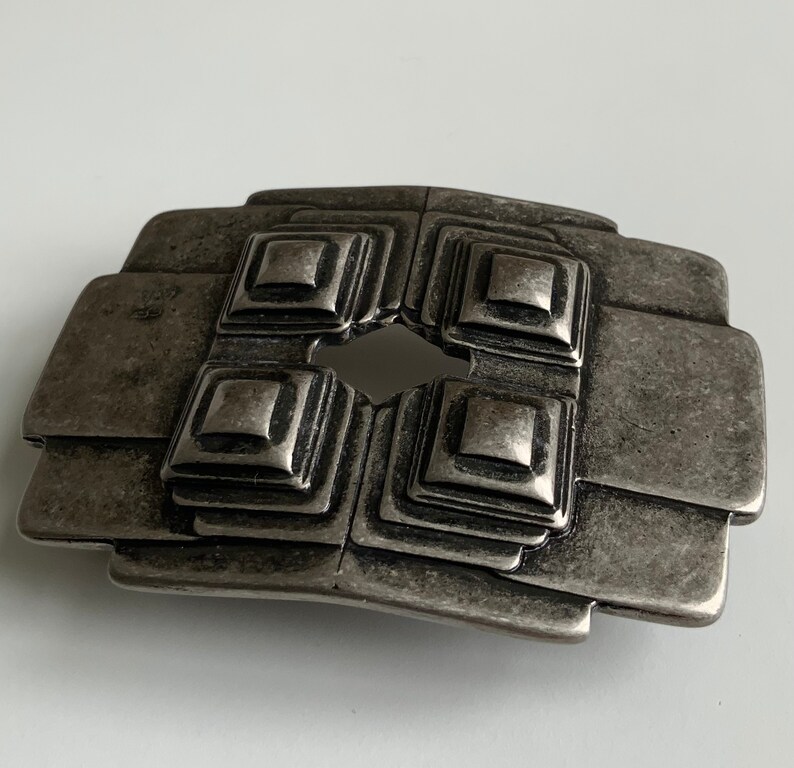 May include: A silver-toned metal belt buckle with a geometric design. The buckle has four square shapes with raised edges and a central hole.