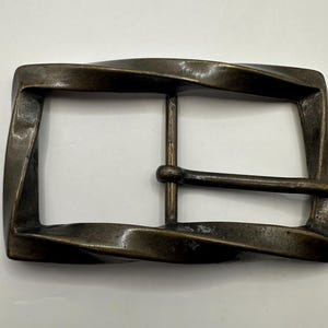 Könnte beinhalten: Eine bronzefarbene Gürtelschnalle mit einem gedrehten, wellenförmigen Design. Die Schnalle hat eine rechteckige Form mit einer glatten, polierten Oberfläche.