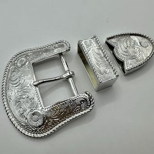 May include: A silver-colored, three-piece belt buckle set. The buckle features intricate floral and scrollwork designs. The set includes a buckle, a belt loop, and a tip, all with a detailed, engraved pattern.