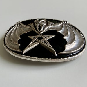 May include: Silver belt buckle with a black enamel background featuring a pentagram and a bat with wings spread out.