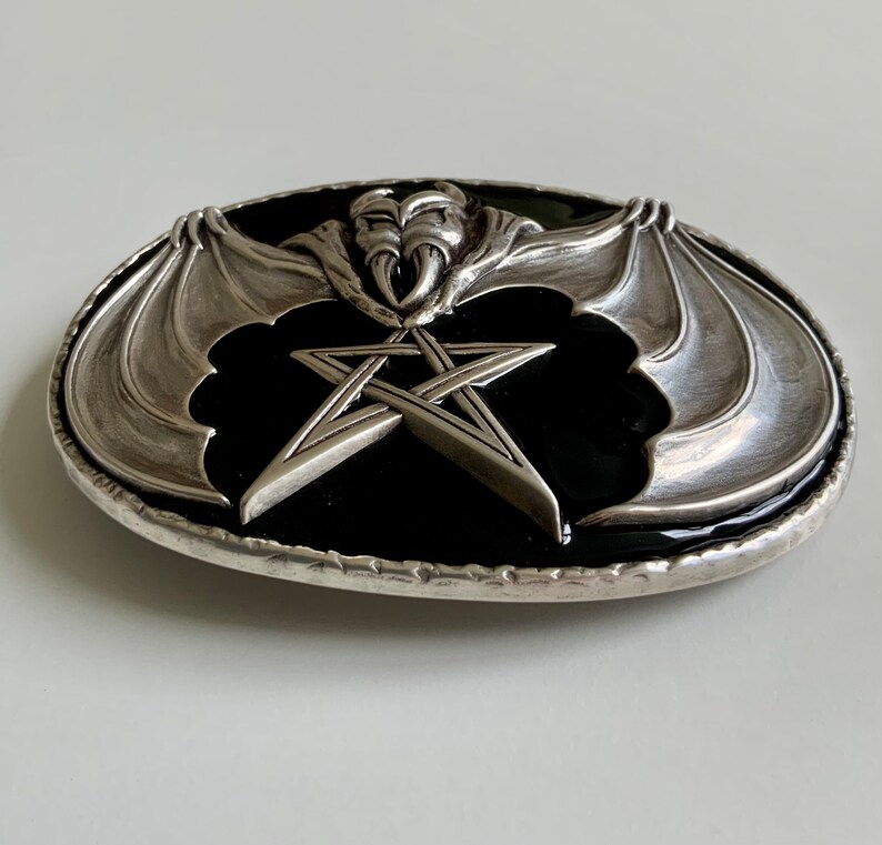 May include: Silver belt buckle with a black enamel background featuring a pentagram and a bat with wings spread out.
