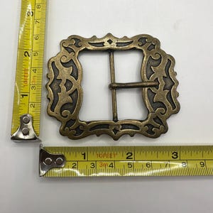 May include: A large, ornate, brass belt buckle with a decorative, filigree design. The buckle is approximately 2 inches wide.