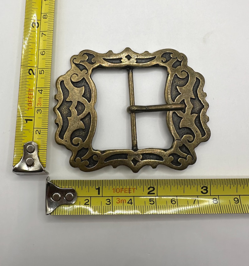 May include: A large, ornate, brass belt buckle with a decorative, filigree design. The buckle is approximately 2 inches wide.