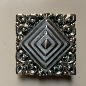 May include: A silver-toned metal square buckle with a decorative, ornate design. The center of the buckle features a raised, diamond-shaped pattern.