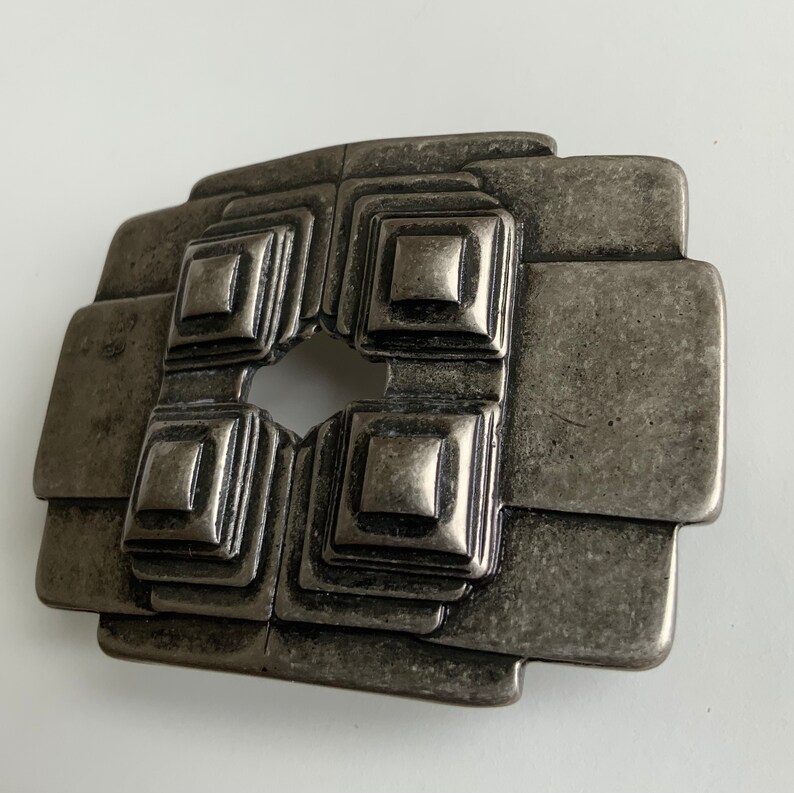 May include: A silver-toned metal belt buckle with a geometric design. The buckle has a square shape with four smaller squares inside, each with a raised center. The buckle has a hole in the center for the belt strap.