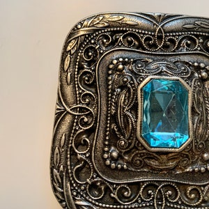 May include: A silver-toned metal buckle with an ornate design and a light blue gemstone in the center.