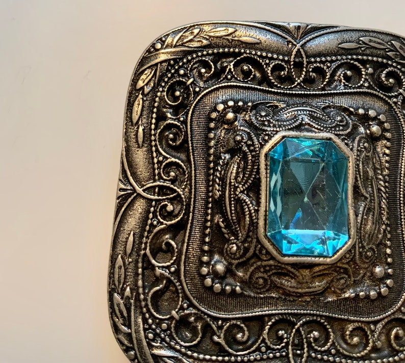 May include: A silver-toned metal buckle with an ornate design and a light blue gemstone in the center.
