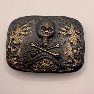 May include: A black metal belt buckle with a gold skull and crossbones design. The buckle has a textured surface and intricate details.