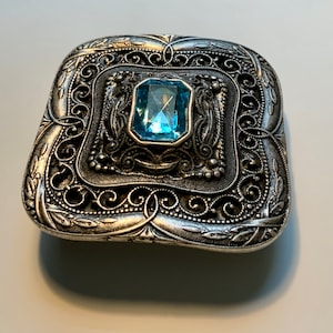 May include: A silver-toned metal brooch with an ornate filigree design and a light blue gemstone in the center.