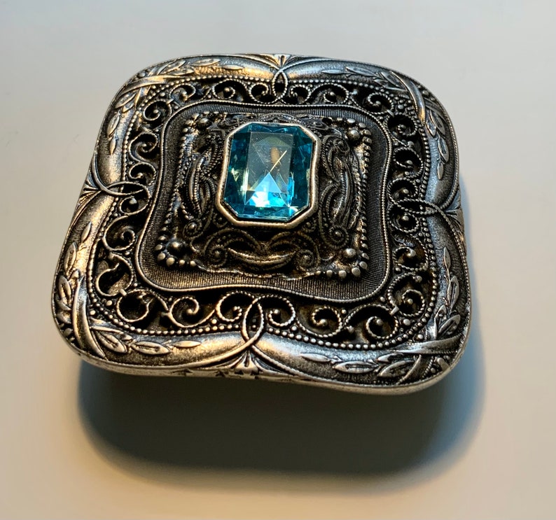 May include: A silver-toned metal brooch with an ornate filigree design and a light blue gemstone in the center.