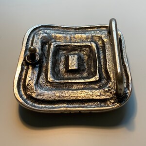 May include: Silver belt buckle with a square design and a textured surface.