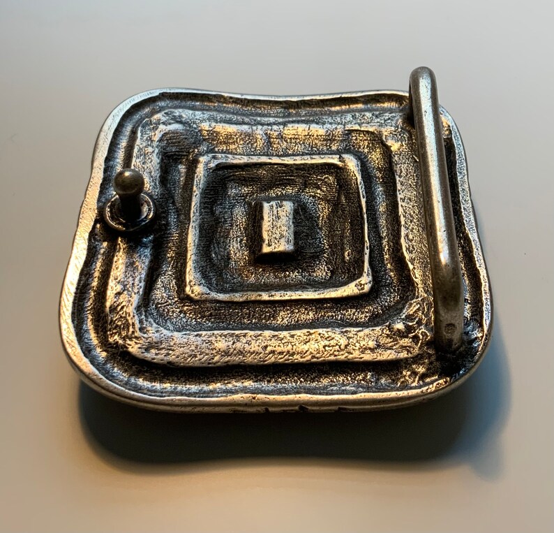 May include: Silver belt buckle with a square design and a textured surface.