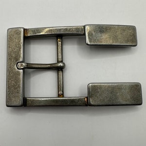 May include: A rectangular, antique silver-colored belt buckle. The buckle has a classic design with a central bar and two rectangular end pieces. The metal has a slightly worn appearance.