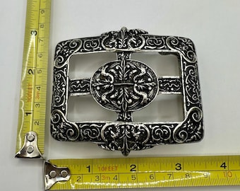 May include: Ornate silver-toned metal belt buckle. The rectangular buckle features an oval center with a cross design and intricate scrollwork. The buckle measures approximately 3.5 inches wide.