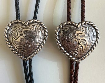 以下が含まれることがあります： ハート型のシルバーペンダントに複雑な彫刻が施された、2本のボロタイ。1つは黒の編み紐、もう1つは茶色の編み紐です。