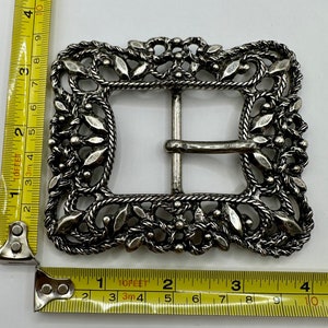 Könnte beinhalten: Silberfarbener Gürtelschnalle aus Metall mit einem filigranen Design aus Blättern und Blumen. Die Schnalle ist etwa 5 cm breit.