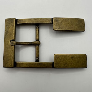 Puede incluir: Hebilla de cinturón rectangular de latón antiguo. La hebilla tiene un diseño clásico con una barra central y dos piezas laterales rectangulares. El metal tiene un aspecto ligeramente desgastado.