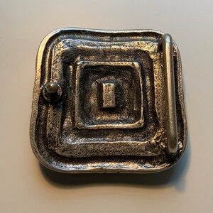 May include: Silver-toned belt buckle with a square design and a textured surface. The buckle has a rectangular opening for a belt.