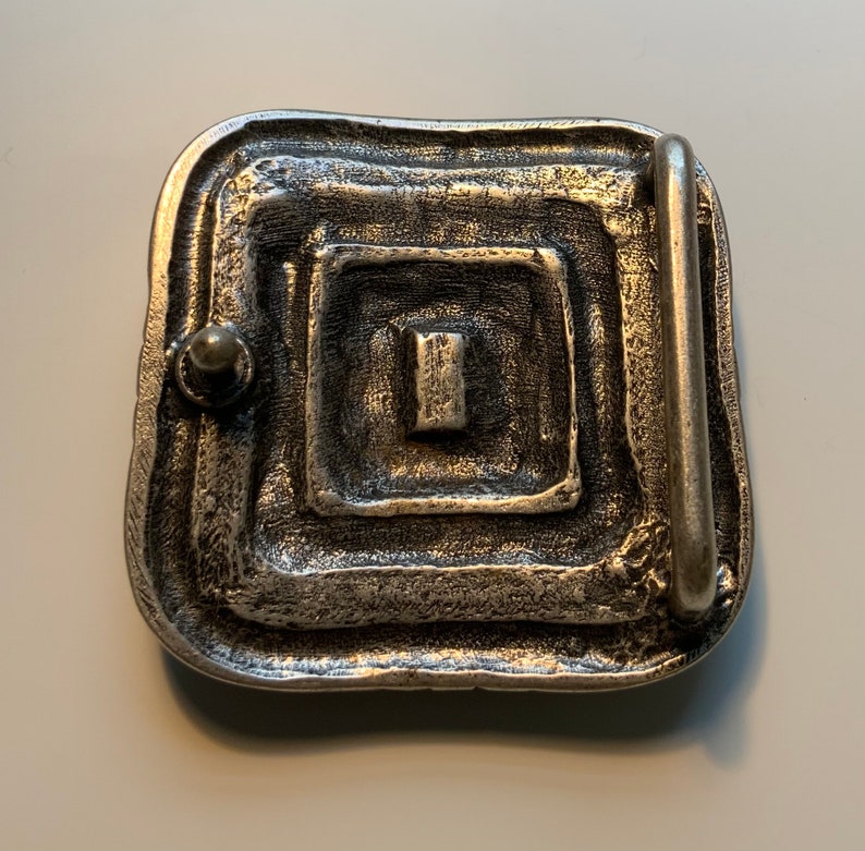 May include: Silver-toned belt buckle with a square design and a textured surface. The buckle has a rectangular opening for a belt.