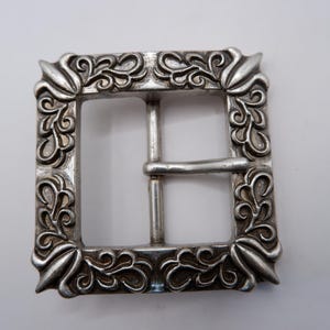 Pode incluir: Uma fivela de cinto quadrada de metal prateado com um padrão de volutas em relevo ao longo da moldura. A fivela inclui um pino central e uma barra horizontal, com elementos decorativos pontiagudos em cada um dos quatro cantos da moldura.