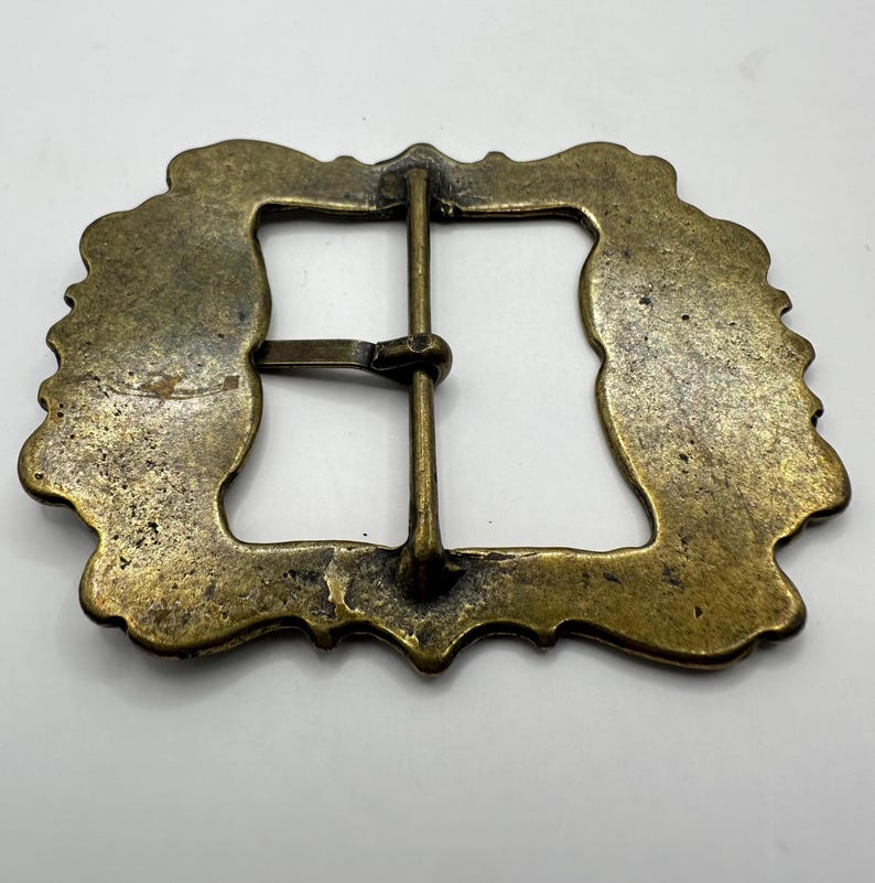 May include: A gold-toned metal belt buckle with an ornate, rectangular frame and a simple, straight bar closure.