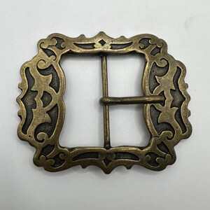 May include: A gold-toned metal belt buckle with an ornate, intricate design. The buckle has a square shape with rounded corners and a detailed, decorative border.