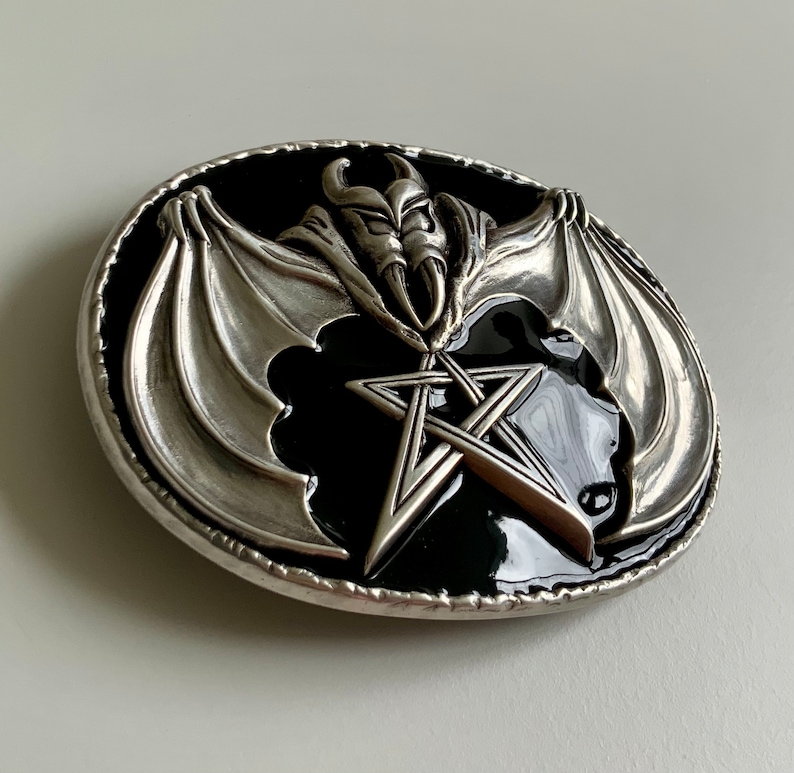 May include: Silver belt buckle with a black enamel background featuring a pentagram and a bat with wings spread out.