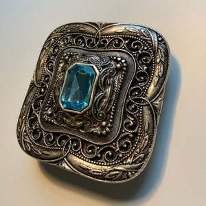May include: A silver-toned, ornate, square-shaped brooch with a light blue gemstone in the center.