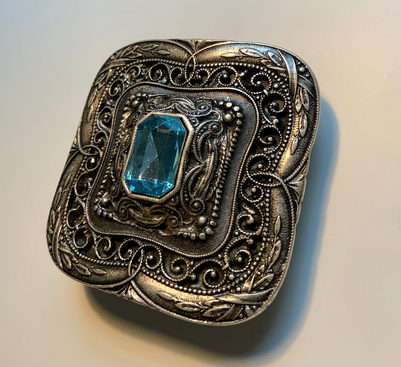 May include: A silver-toned, ornate, square-shaped brooch with a light blue gemstone in the center.