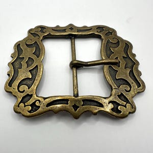 May include: A gold-toned metal belt buckle with an ornate, intricate design. The buckle has a square shape with a decorative border.