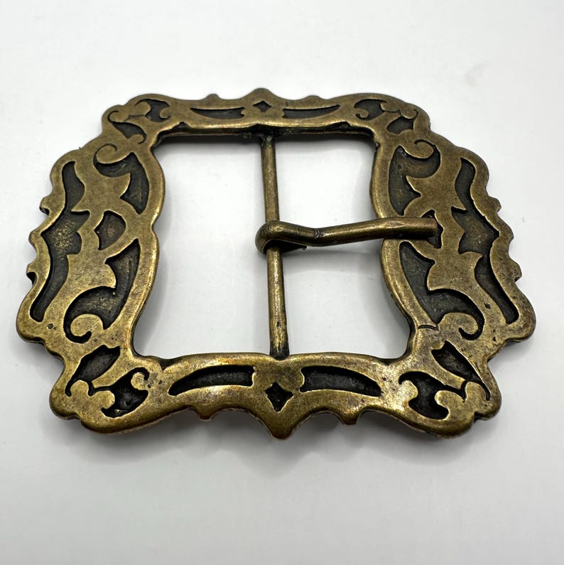 May include: A gold-toned metal belt buckle with an ornate, intricate design. The buckle has a square shape with a decorative border.