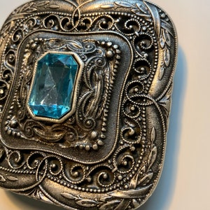 May include: A silver-toned metal brooch with an ornate design featuring a large, light blue gemstone in the center.