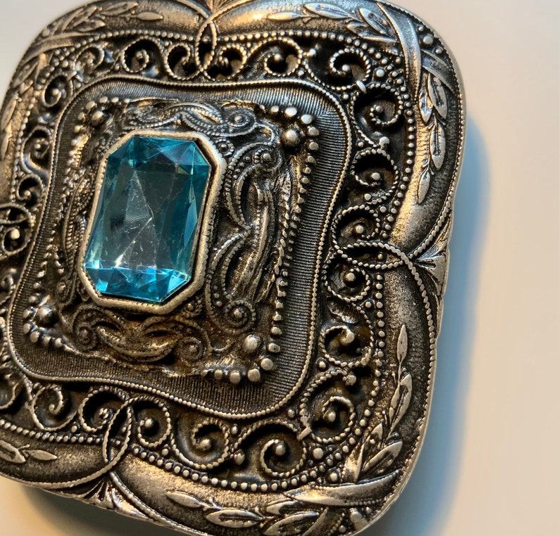 May include: A silver-toned metal brooch with an ornate design featuring a large, light blue gemstone in the center.