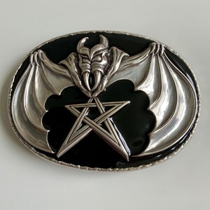 May include: A silver belt buckle with a black enamel background. The buckle features a bat with wings spread wide and a five-pointed star in the center.