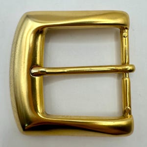 May include: A gold-colored metal belt buckle with a rectangular shape and rounded corners. The buckle has a single bar and a prong for securing a belt. The metal has a shiny finish.