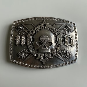 May include: Silver belt buckle with a skull design and the Harley Davidson logo. The buckle is decorated with rhinestones and has the word "H.O.G." on either side of the skull.