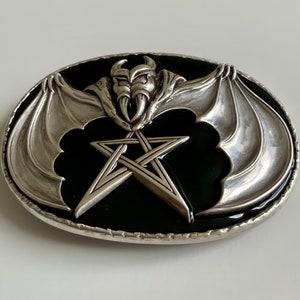 May include: A silver belt buckle with a black enamel background. The buckle features a pentagram with a bat with wings spread out on either side of the star.