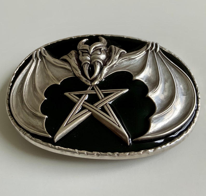 May include: A silver belt buckle with a black enamel background. The buckle features a pentagram with a bat with wings spread out on either side of the star.